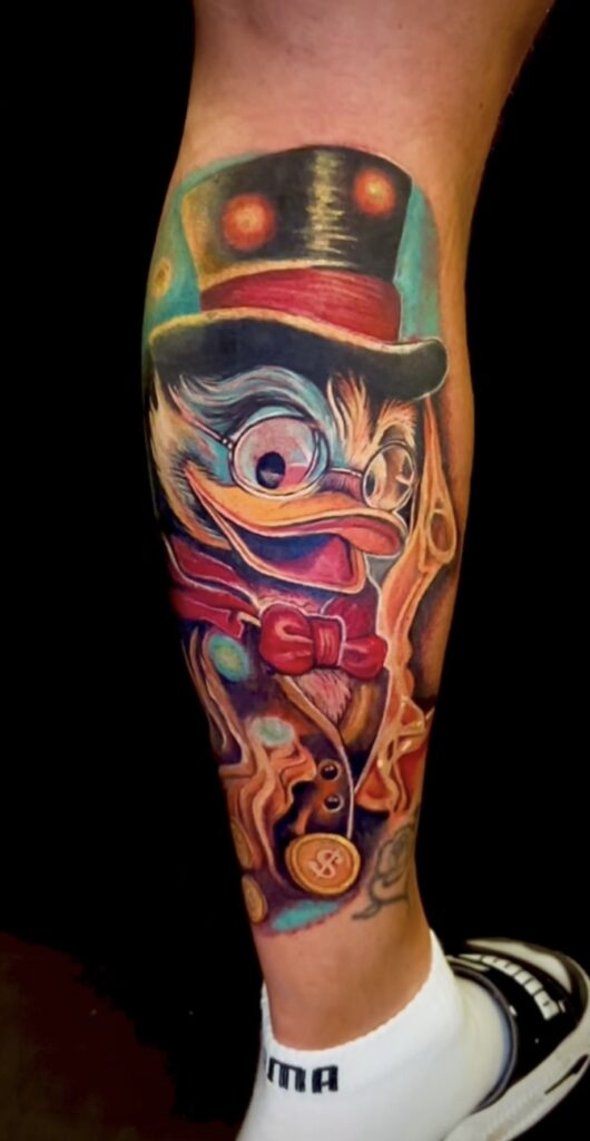 "Een kleurrijke, illustratieve tattoo van Dagobert Duck (Scrooge McDuck) op een onderbeen. De tattoo is rijk aan details, met Dagobert die een hoge hoed, bril en een rode strik draagt, omringd door gouden munten. Gezet door Sanne van Santji Art op de Sint-Niklaas Tattoo Expo.