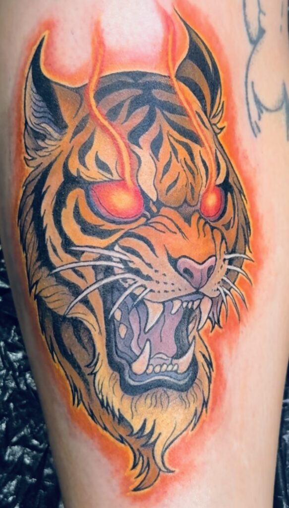 Een krachtige tattoo van een brullende tijgerkop in neo-traditional stijl. De tijger heeft oplichtende rode ogen waaruit vlammen opstijgen, omlijst door een vurige oranje gloed. Gezet door Sanne van Santji Art op de Sint-Niklaas Tattoo Expo.