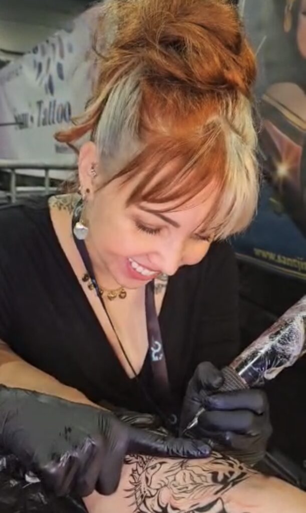 Een actiefoto van tattoo-artiest Sanne (Santji Art) die glimlachend een tatoeage zet tijdens de Sint-Niklaas Tattoo Expo. Ze heeft opvallend rood met blond haar en draagt zwarte handschoenen terwijl ze met precisie aan een ontwerp op een arm werkt.