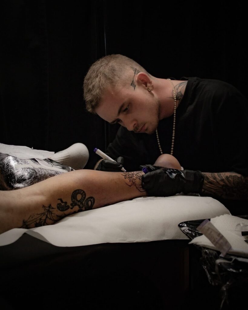 Sfeerbeeld van tattoo-artiest Brett (Tattoossenon) op de Sint-Niklaas Tattoo Expo. Hij is geconcentreerd bezig met het voortekenen van een ontwerp op het been van een klant met markers (freehand techniek), wat zijn artistieke proces prachtig in beeld brengt.