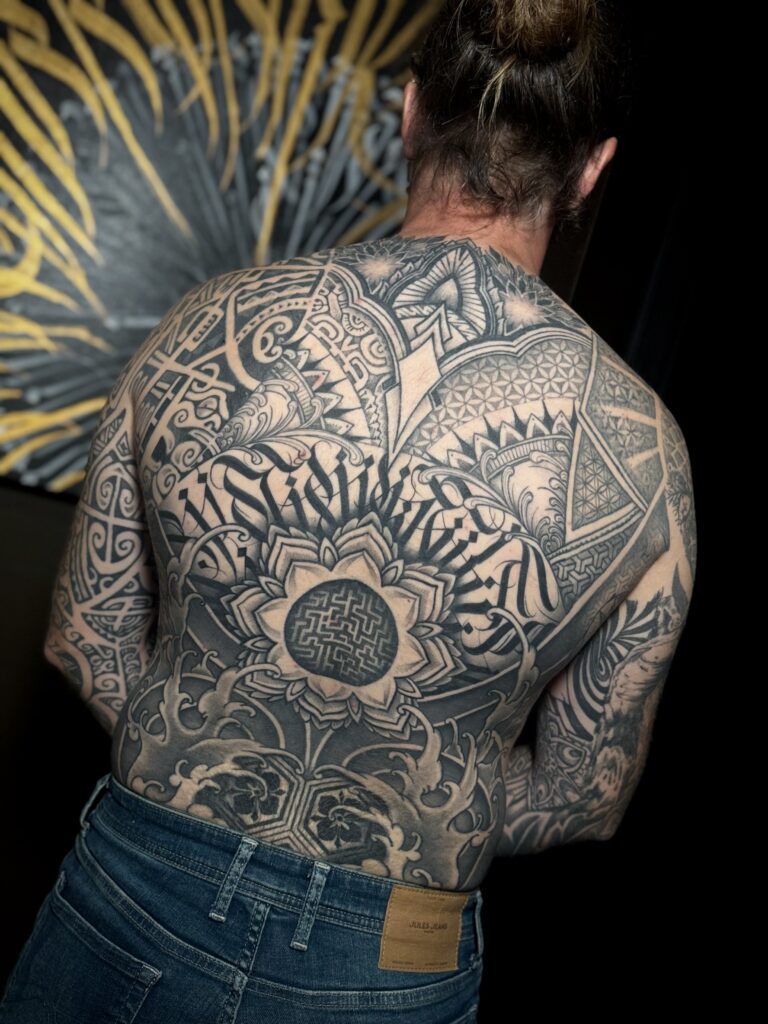 Een indrukwekkend, volledig rugstuk in blackwork stijl door Brett (Tattoossenon). Het ontwerp is een complexe compositie van geometrische patronen (sacred geometry), mandala's en krachtige calligrafische letters die de hele rug bedekken. Gefotografeerd op de Sint-Niklaas Tattoo Expo.