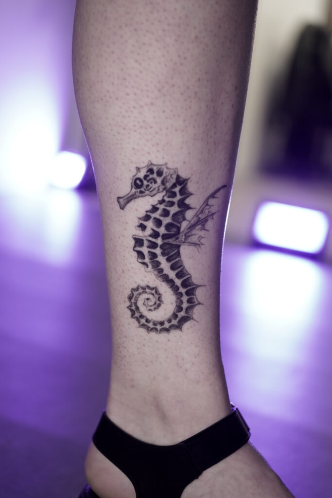 Een elegante fineline tattoo van een zeepaardje op een enkel. Het zeepaardje is zeer gedetailleerd uitgewerkt met organische texturen en fijne vinnen, geplaatst tegen een sfeervolle paarse achtergrondverlichting. Werk van Naviia (Be Inkspired Tattoo) op de Sint-Niklaas Tattoo Expo.