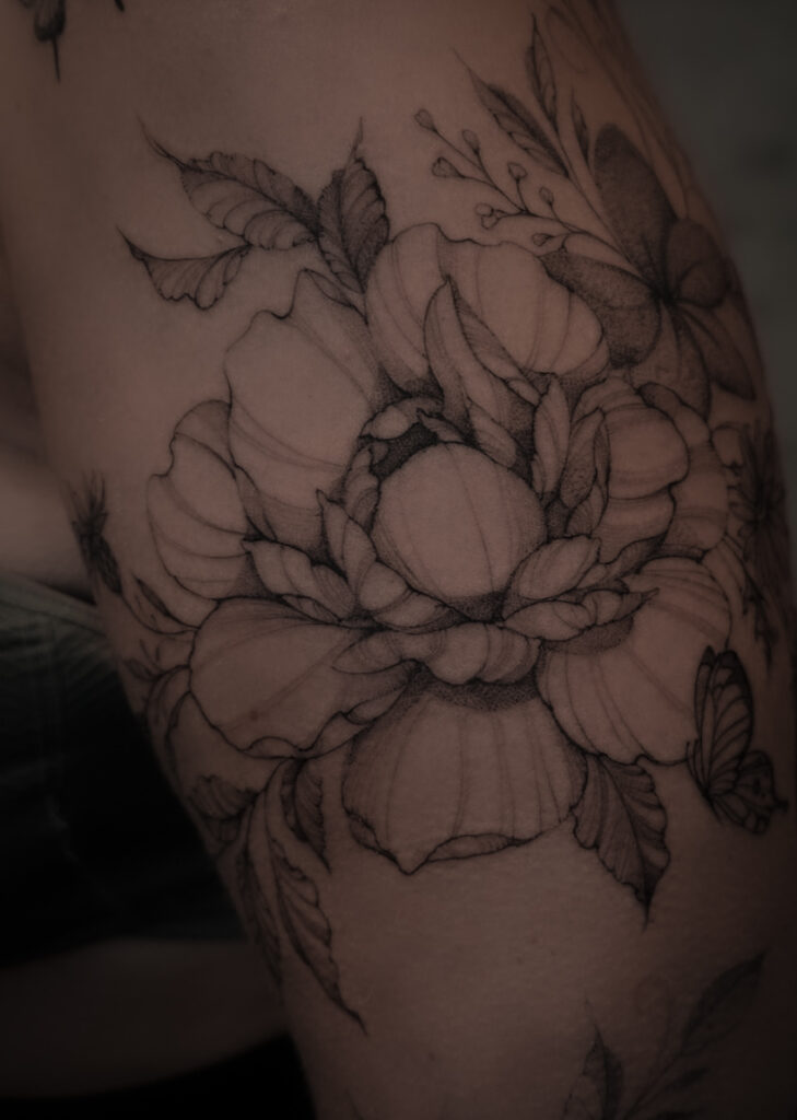 Een close-up van een botanische tattoo die een pioenroos in volle bloei toont, omringd door bladeren en een kleine vlinder. Het ontwerp is uitgevoerd in een subtiele black and grey stijl met veel oog voor detail. Werk van Sasha Hemelaer Tattoo op de Sint-Niklaas Tattoo Expo.