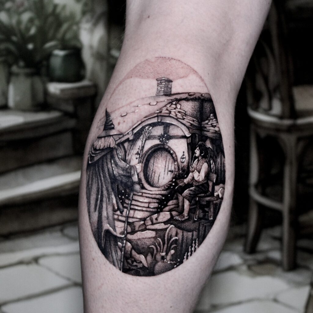 Een gedetailleerde, ronde blackwork tattoo die de iconische scène bij Bag End uit The Hobbit weergeeft. Men ziet Gandalf de Grijze bij de ronde deur van de hobbitwoning, terwijl Bilbo Balbaas op een bankje toekijkt. De tattoo is rijk aan texturen zoals steen, hout en planten. Gezet door Jana van Jana Jacques Tattoo op de Sint-Niklaas Tattoo Expo.