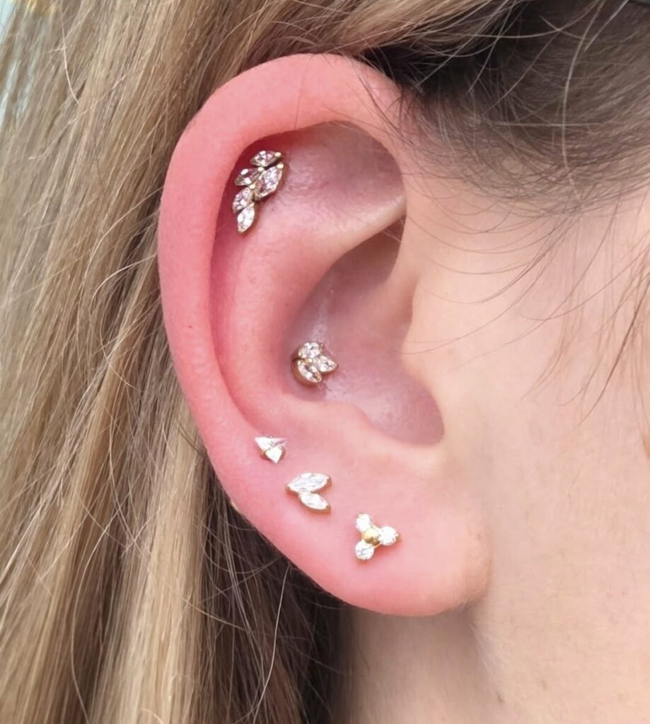 Een verfijnde set piercings in de oorlel en het kraakbeen, uitgevoerd in goud met marquise-geslepen kristallen die lijken op botanische blaadjes. Een harmonieuze en luxe uitstraling, verzorgd door Jolene (Piercexpression) op de Sint-Niklaas Tattoo Expo.