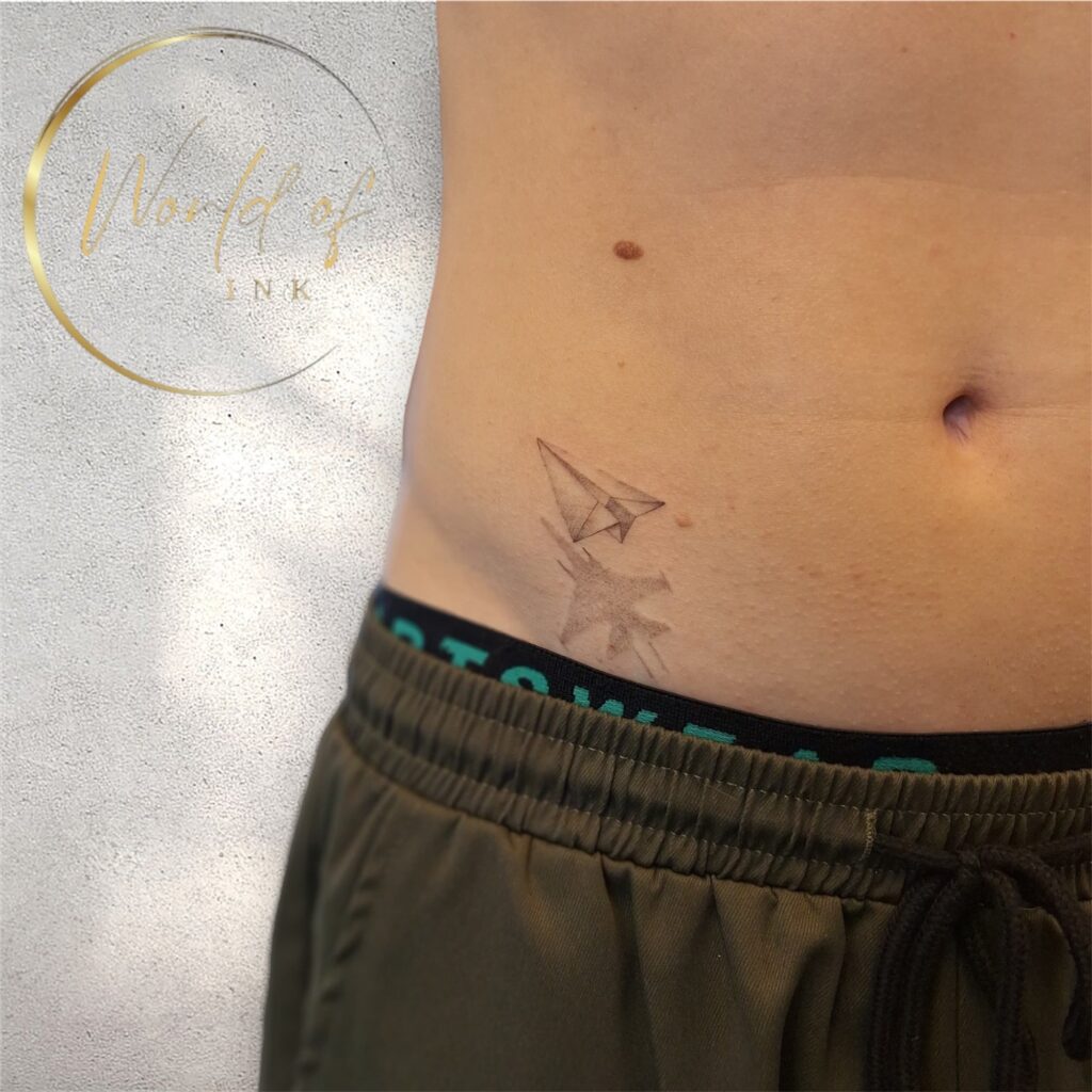 Een subtiele fineline tattoo op de heup van een gevouwen papieren vliegtuigje dat een schaduw werpt in de vorm van een gevechtsvliegtuig. De tattoo is gezet met zeer dunne lijnen en zachte schaduwwerking. Gezet door Angelika van Go Ink Tattoos tijdens de Sint-Niklaas Tattoo Expo.