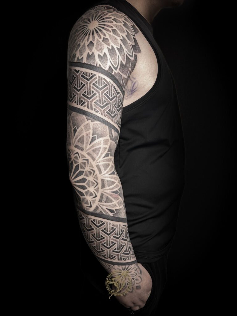 Een volledige arm-sleeve in geometrische blackwork-stijl. Het ontwerp bevat een grote mandala op de schouder en bovenarm, afgewisseld met strakke banden van complexe, 3D-achtige patronen en fijn dotwork. Werk van Zoraya (Ink About It Tattoo) op de Sint-Niklaas Tattoo Expo.