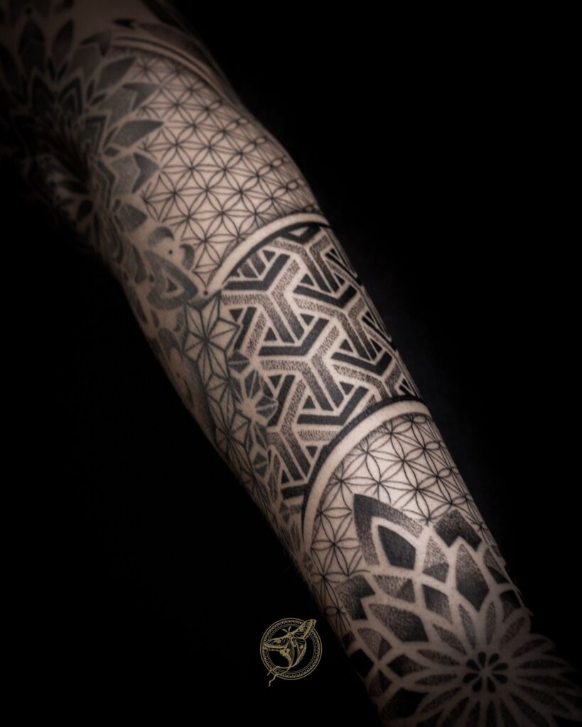 Een close-up van een tatoeage op de arm die verschillende geometrische technieken combineert. Te zien zijn patronen van de 'Flower of Life', mandala-achtige bloemvormen en scherpe architecturale lijnen, allemaal uitgevoerd met uiterst nauwkeurig dotwork en diepe zwarttinten. Gezet door Zoraya van Ink About It Tattoo op de Sint-Niklaas Tattoo Expo.