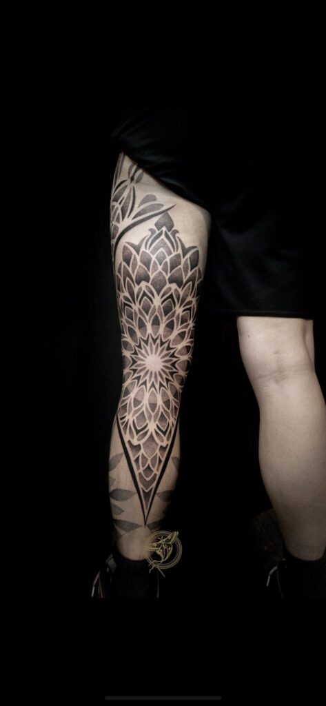 Een grootschalige, verticale mandala-tatoeage op de achterzijde van het bovenbeen en de knieholte. Het ontwerp is puntig en symmetrisch, opgebouwd uit schaduwrijke bloembladen en fijn dotwork, wat zorgt voor een krachtig visueel effect. Vakmanschap van Zoraya (Ink About It Tattoo) op de Sint-Niklaas Tattoo Expo.