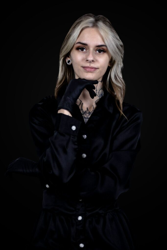 Een professioneel portret van tattoo-artiest Zoraya (Ink About It Tattoo) tegen een zwarte achtergrond. Ze draagt een zwart satijnen overhemd en zwarte handschoenen, waarbij haar hand onder haar kin rust. Haar eigen nektattoos en piercings zijn zichtbaar, wat haar artistieke identiteit benadrukt.