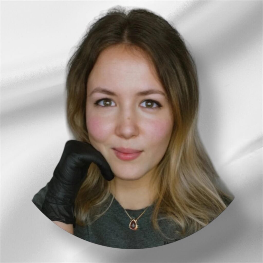 Een portret van tattoo-artiest Angelika (Go Ink Tattoos) in een ronde uitsnede tegen een lichte achtergrond. Ze heeft blond haar, draagt een donkergrijs shirt met een ketting en een zwarte tatoeagehandschoen aan haar rechterhand die haar kin ondersteunt.