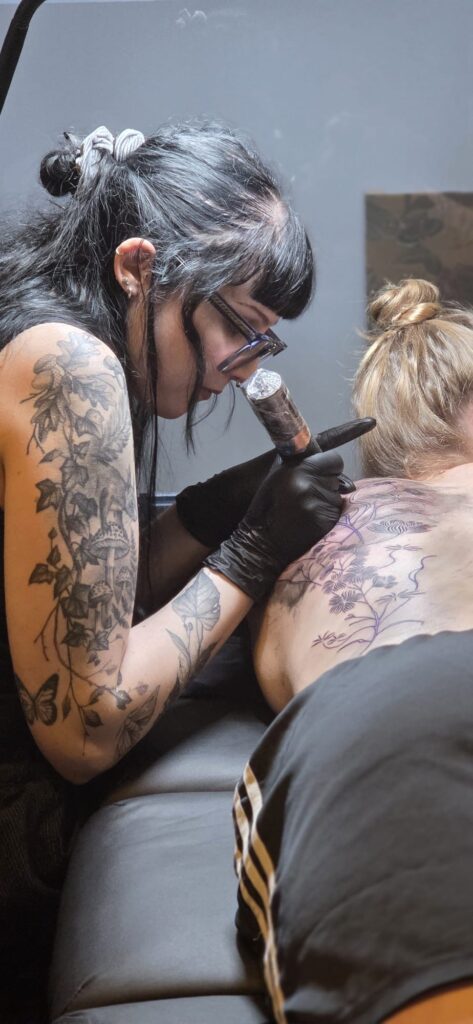 Een actiefoto van tattoo-artiest Morgan (Morgan le Fay) die geconcentreerd een grote rugtattoo zet op de Sint-Niklaas Tattoo Expo. Ze draagt een bril en zwarte handschoenen, terwijl haar eigen gedetailleerde armtattoos van planten en paddenstoelen goed zichtbaar zijn.