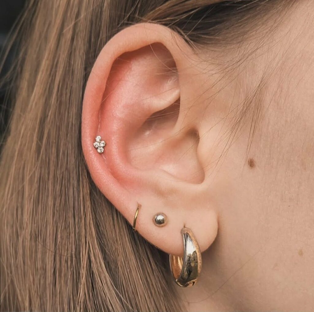 Close-up van een oorcuratie in goudkleurige tinten. Het oor bevat een helix-piercing met kristallen in bloemvorm, een minimalistische ring en een bolletje in de oorschelp, en een grotere gouden hoop in de oorlel. Gepierced door Jolene van Piercexpression tijdens de Sint-Niklaas Tattoo Expo.