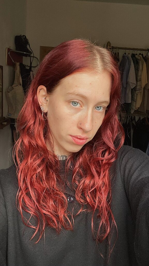 Portretfoto van tattoo-artiest Wietske (Tatties Wietske) op de Sint-Niklaas Tattoo Expo. Ze heeft golvend rood haar en draagt een zwarte top. Verschillende piercings zijn zichtbaar in haar oren en neus, evenals een tattoo in haar nek.