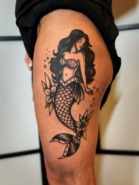 Een klassieke blackwork tattoo van een zeemeermin in traditional stijl op een bovenbeen. De zeemeermin heeft lang, golvend zwart haar en een gedetailleerde geschubde staart, omringd door bloemen en kleine luchtbellen. Gezet door Joe van Joe Crooked Tattoo op de Sint-Niklaas Tattoo Expo.