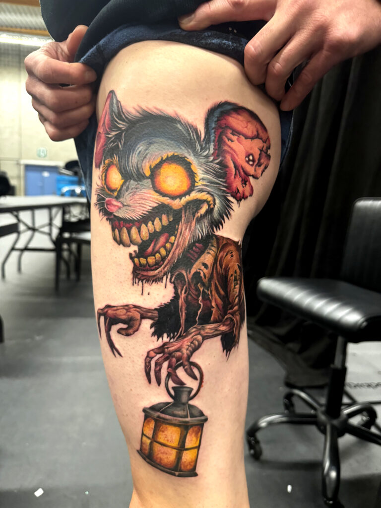 Een indrukwekkende en levendige tattoo van een monsterlijke muis in horror-stijl op een bovenbeen. Het personage heeft gloeiende oranje ogen, een grote grijns met scherpe tanden en houdt een brandende lantaarn vast. De tattoo valt op door het sterke kleurcontrast en de gedetailleerde vacht. Gezet door Sabrina van Scarlett Ink op de Sint-Niklaas Tattoo Expo.