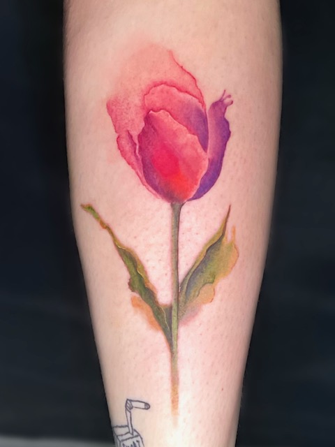 Een verfijnde watercolor tattoo van een tulp op een onderarm, uitgevoerd in vloeiende roze en paarse tinten met zachte groene bladeren. Gezet door artiest Annet van Ink Therapy tijdens de Sint-Niklaas Tattoo Expo.