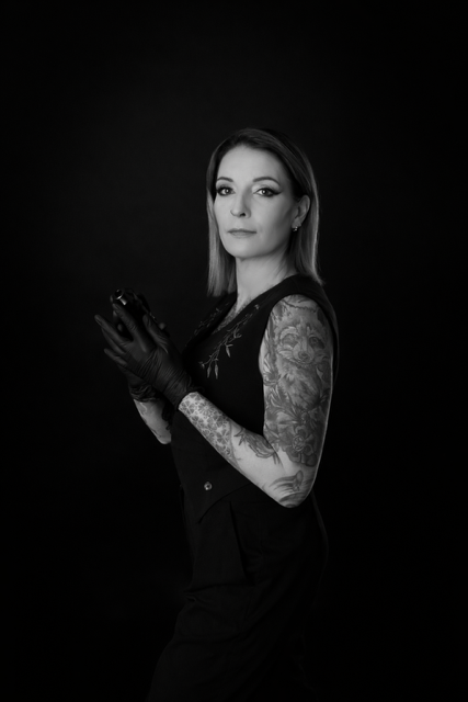 Een stijlvolle zwart-wit studiofoto van tattoo-artiest Sofie (Jedaloe Tattoo). Ze draagt een zwart gilet en heeft opvallende tattoos op haar armen, waaronder een wasbeer en florale patronen. Ze kijkt rustig in de camera tegen een donkere achtergrond.