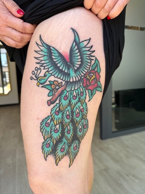 Een kleurrijke traditional tattoo van een pauw in volle vlucht op een bovenbeen. Het ontwerp gebruikt klassieke tinten zoals groen, rood en geel, en is versierd met een roze roos aan de zijkant. Werk van Joe (Joe Crooked Tattoo) op de Sint-Niklaas Tattoo Expo.