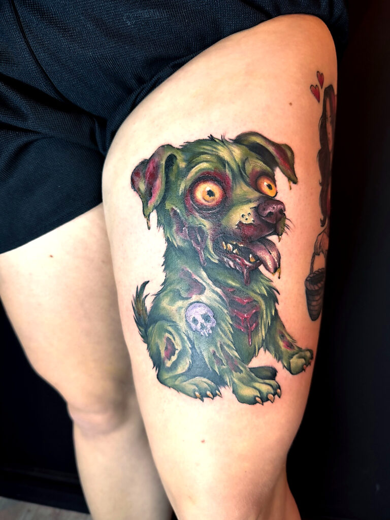 Een kleurrijke en gedetailleerde horror-stijl tattoo van een zombie-hondje op een bovenbeen. De hond heeft een groene huid, uitpuilende gele ogen en diverse wonden waar bloed en bot zichtbaar zijn. Op de heup van de hond staat een klein schedeltje getatoeëerd. Werk van Sabrina (Scarlett Ink) op de Sint-Niklaas Tattoo Expo.