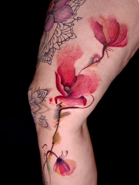 Artistieke watercolor tattoo van rode klaprozen op een been, gecombineerd met subtiele abstracte kleurspatten en zwarte geometrische mandala-details op de achtergrond. Werk van Annet (Ink Therapy) op de Sint-Niklaas Tattoo Expo.