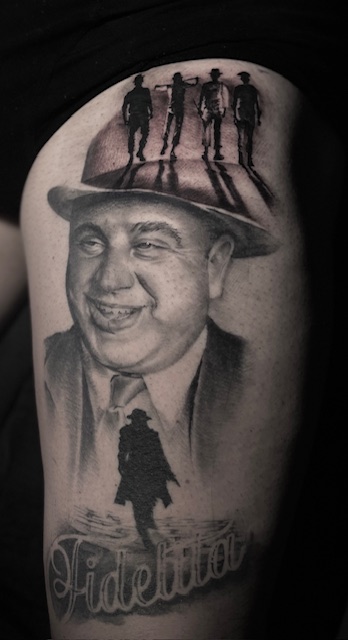 Een gedetailleerde black and grey tattoo op een been, met een realistisch portret van een lachende man in een pak en hoed. Op de rand van de hoed lopen silhouetten van figuren uit 'A Clockwork Orange', en onderaan staat een donker silhouet van een man in een lange jas bij het woord 'Fedeltà'. Gezet door Sofie van Jedaloe Tattoo op de Sint-Niklaas Tattoo Expo.