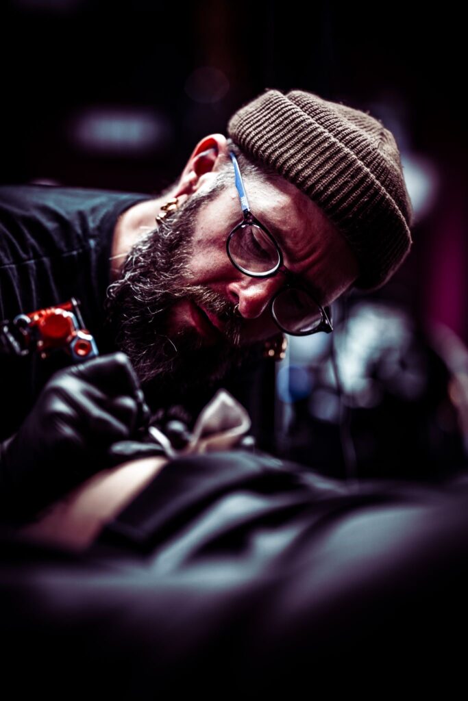 Een sfeervolle, artistieke actiefoto van tattoo-artiest Joe (Joe Crooked Tattoo) op de Sint-Niklaas Tattoo Expo. Joe is geconcentreerd aan het tatoeëren en draagt een bruine muts, een bril en zwarte handschoenen. De foto heeft een diep contrast en een warme, professionele uitstraling.