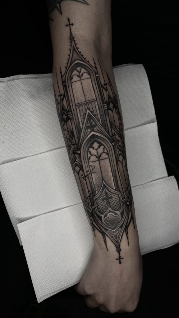 Een grootschalige, verticale tattoo op een onderarm die de gedetailleerde architectuur van een gotische kathedraal weergeeft, inclusief spitsbogen, glas-in-lood ramen en kruisbeelden. De tattoo is gezet met strakke zwarte lijnen en zacht schaduwwerk. Gezet door Kim van Transylveinia Tattoo tijdens de Sint-Niklaas Tattoo Expo.