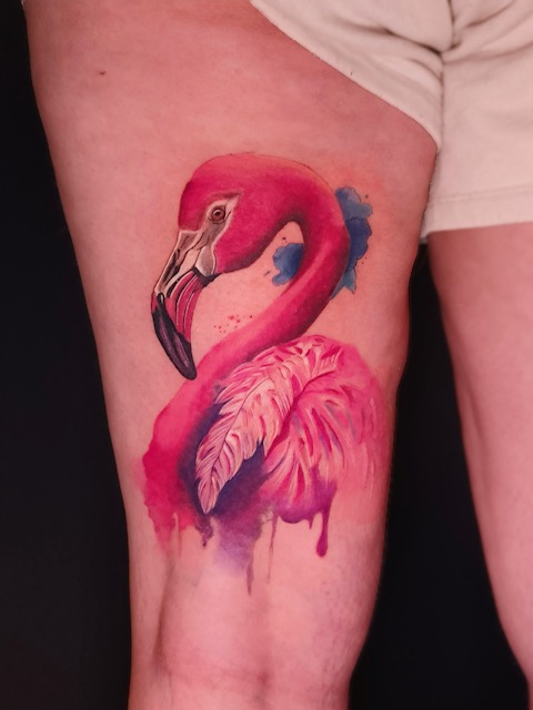 Kleurrijke tattoo van een flamingo in watercolor-stijl op een bovenbeen. De vogel is gedetailleerd uitgewerkt in dieproze en paars met contrasterende blauwe inktspatten onderaan. Gezet door Annet van Ink Therapy op de Sint-Niklaas Tattoo Expo.