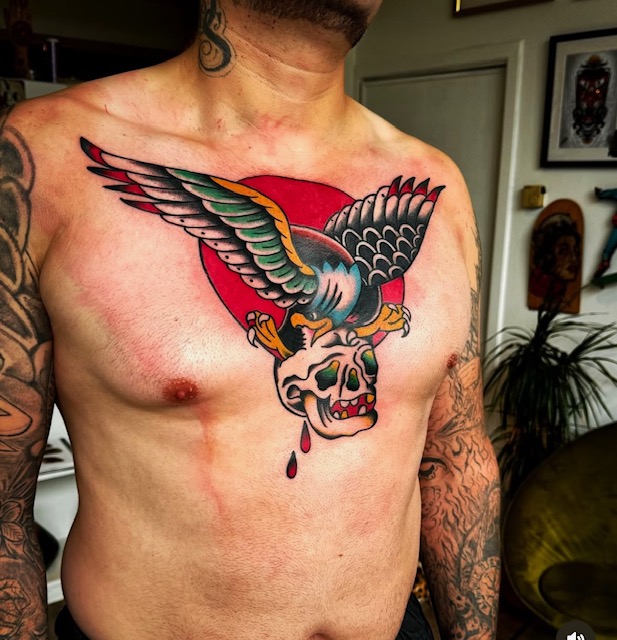 Een krachtig traditional borststuk van een arend die zijn klauwen in een schedel zet, tegen de achtergrond van een grote rode zon. De arend is uitgevoerd in de klassieke kleurencombinatie van zwart, groen en geel. Gezet door Joe van Joe Crooked Tattoo tijdens de Sint-Niklaas Tattoo Expo.