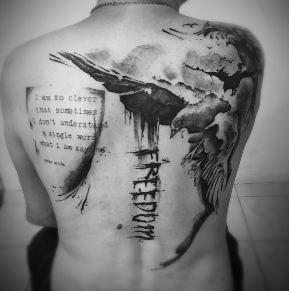 Een artistiek black and grey rugstuk in een dromerige watercolor-stijl. Het ontwerp toont een grote vogel in vlucht die overgaat in abstracte inktspatten. Aan de linkerzijde staat een citaat van Oscar Wilde in een getypt lettertype, en centraal over de ruggengraat loopt het woord FREEDOM in krachtige, verticale letters. Werk van Kaly (Tattoo Kaly) op de Sint-Niklaas Tattoo Expo.