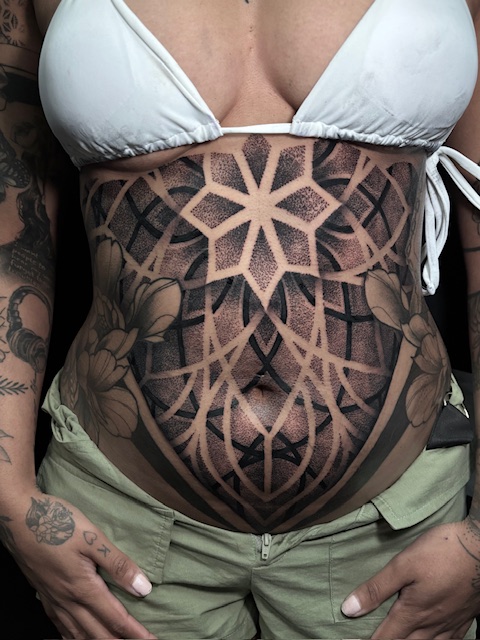 Een grootschalige, symmetrische geometrische tattoo op de buik. Het ontwerp bestaat uit een centrale ster vormgegeven met fijn dotwork, omringd door complexe mandalapatronen en subtiele bloemdetails aan de zijkanten. Vakmanschap van Sofie (Jedaloe Tattoo) op de Sint-Niklaas Tattoo Expo.
