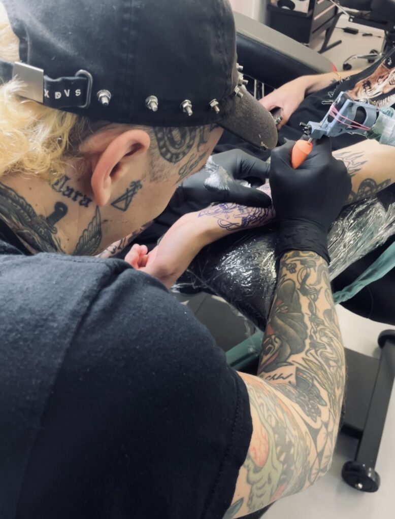Een actiefoto van tattoo-artiest Barrie (Black Rain Tattoo) op de Sint-Niklaas Tattoo Expo. Hij draagt een zwarte pet met spikes en is geconcentreerd bezig met het zetten van een tattoo op een onderarm. Zijn eigen armen en nek zijn voorzien van uitgebreide black and grey tattoos.