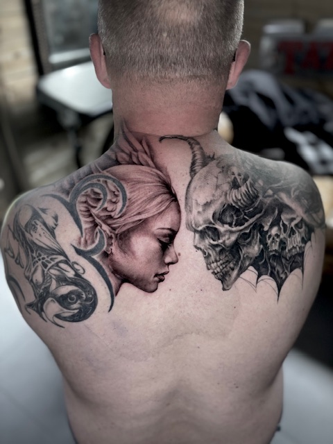 Een surrealistische black and grey tattoo op de bovenrug en nek. De compositie toont het profiel van een vrouwelijk gezicht dat recht tegenover een gedetailleerde schedel met hoorns staat, geflankeerd door abstracte en duistere elementen. Gezet door Sofie van Jedaloe Tattoo op de Sint-Niklaas Tattoo Expo.