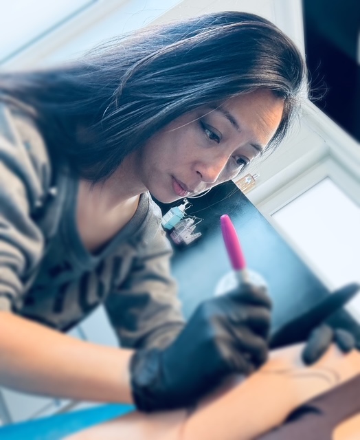 Een sfeervolle foto van tattoo-artiest Kaly (Tattoo Kaly) die geconcentreerd bezig is met een ontwerp. Ze draagt zwarte handschoenen en heeft een roze marker in haar hand, waarmee ze waarschijnlijk een freehand ontwerp op de huid uittekent. Haar focus en vakmanschap zijn duidelijk zichtbaar tijdens de Sint-Niklaas Tattoo Expo.