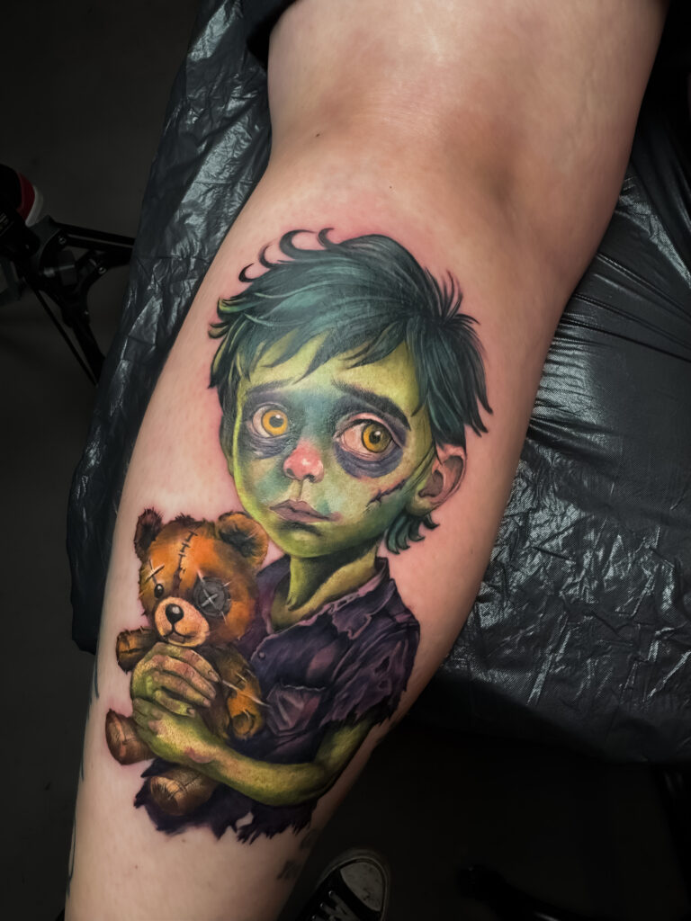 Een aangrijpende en uiterst gedetailleerde horror-tattoo van een jongetje als zombie op een bovenbeen. Het kind heeft een groenachtige huid, diepe kringen onder zijn ogen en houdt een kapotte teddybeer vast. De tattoo toont een hoog niveau van realisme in de texturen en kleurovergangen. Vakmanschap van Sabrina (Scarlett Ink) tijdens de Sint-Niklaas Tattoo Expo.