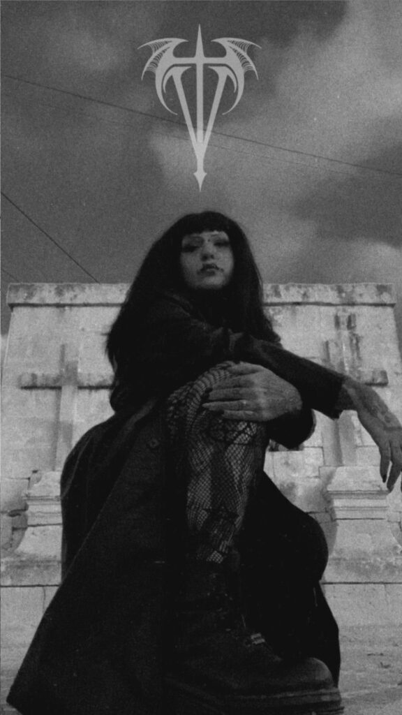 Een sfeervolle zwart-wit foto van tattoo-artiest Kim (Transylveinia Tattoo) voor een gotische stenen achtergrond met kruisen. Ze draagt een donkere outfit en netpanty, passend bij de esthetiek van haar werk. Boven haar hoofd zweeft haar logo in een grafische stijl.