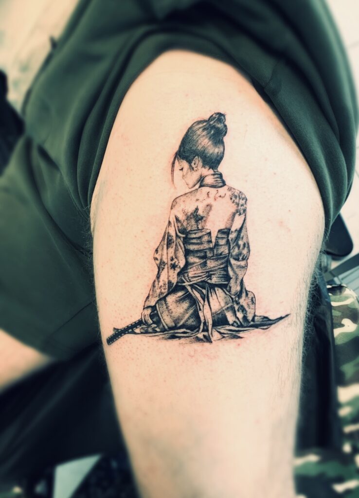 Een uiterst gedetailleerde black and grey tattoo op een bovenarm van een zittende vrouwelijke samoerai, gezien vanaf de achterkant. Ze draagt een traditionele kimono en heeft een zwaard (katana) naast zich liggen. De tattoo valt op door het fijne schaduwwerk en de realistische weergave van de stoffen. Werk van Kaly (Tattoo Kaly) tijdens de Sint-Niklaas Tattoo Expo.