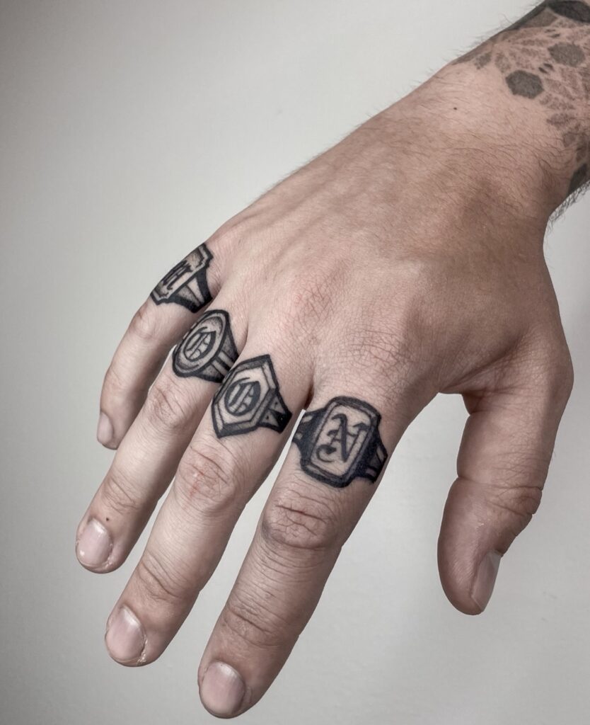 Een close-up van een hand met vier zwarte tattoos op de knokkels in de vorm van klassieke zegelringen. Elke ring bevat een andere letter in een sierlijk lettertype. Werk van Barrie (Black Rain Tattoo) op de Sint-Niklaas Tattoo Expo.