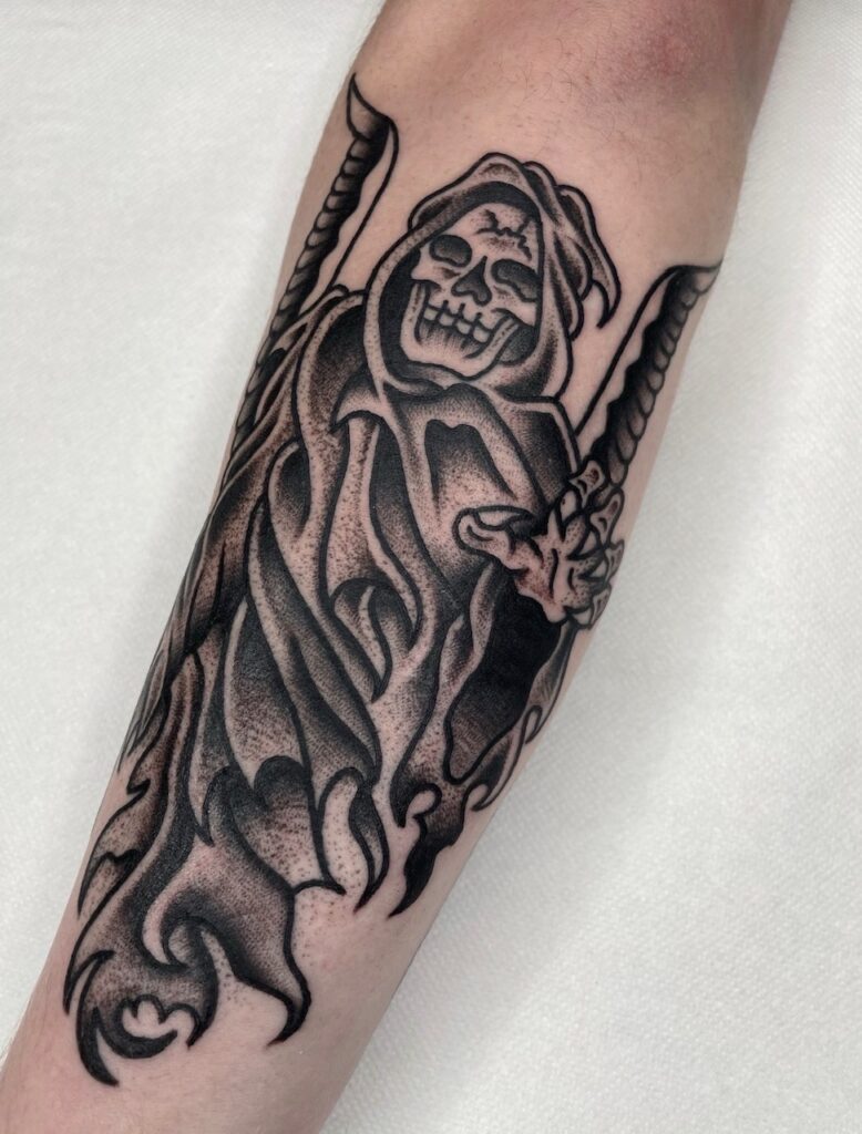Een indrukwekkende black and grey tattoo van een lachende Magere Hein (Grim Reaper) met een zeis. De tattoo kenmerkt zich door stevige zwarte lijnen en zacht dotwork-schaduwwerk op een onderarm. Werk van Barrie (Black Rain Tattoo) tijdens de Sint-Niklaas Tattoo Expo.
