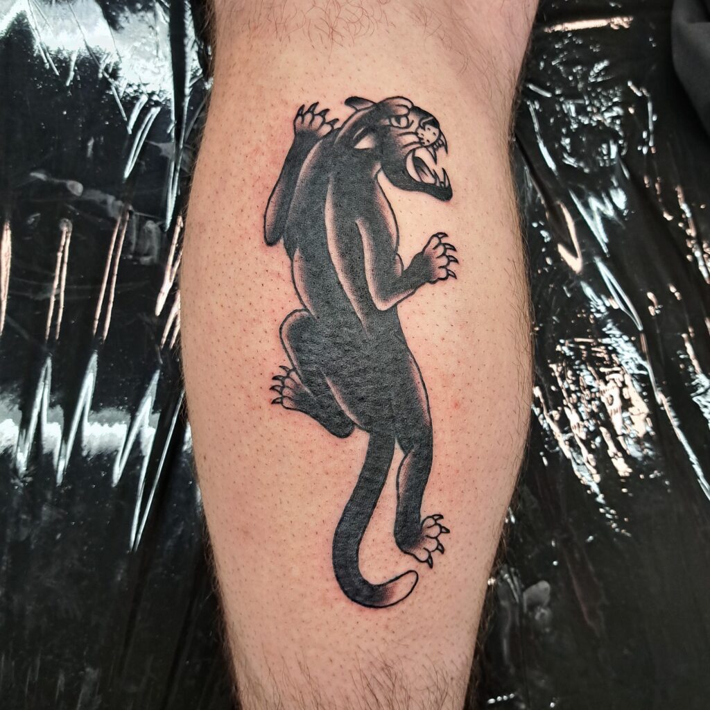 Een krachtige blackwork tattoo van een panter in een klimmende houding op een been. De panter is afgebeeld met ontblote tanden en scherpe klauwen, kenmerkend voor de traditionele tattoo-stijl. Werk van Wietske (Tatties Wietske) op de Sint-Niklaas Tattoo Expo.