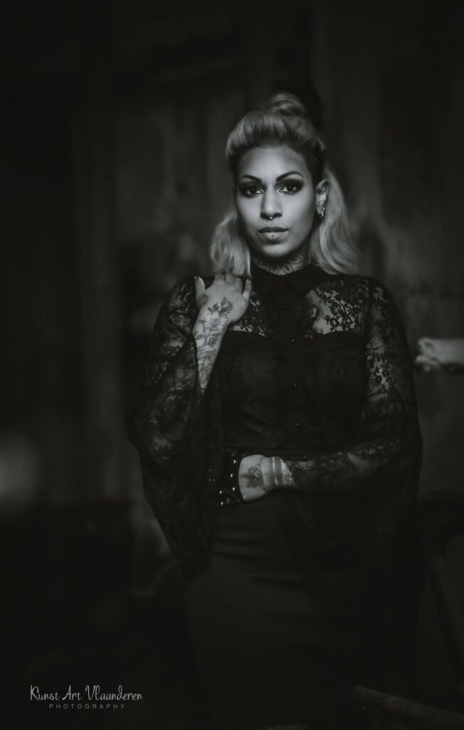 Een sfeervol zwart-wit portret van piercer Jolene (Piercexpression) op de Sint-Niklaas Tattoo Expo. Ze draagt een zwarte kanten top, heeft opvallende hand- en nektattoos, en kijkt met een krachtige blik in de camera.