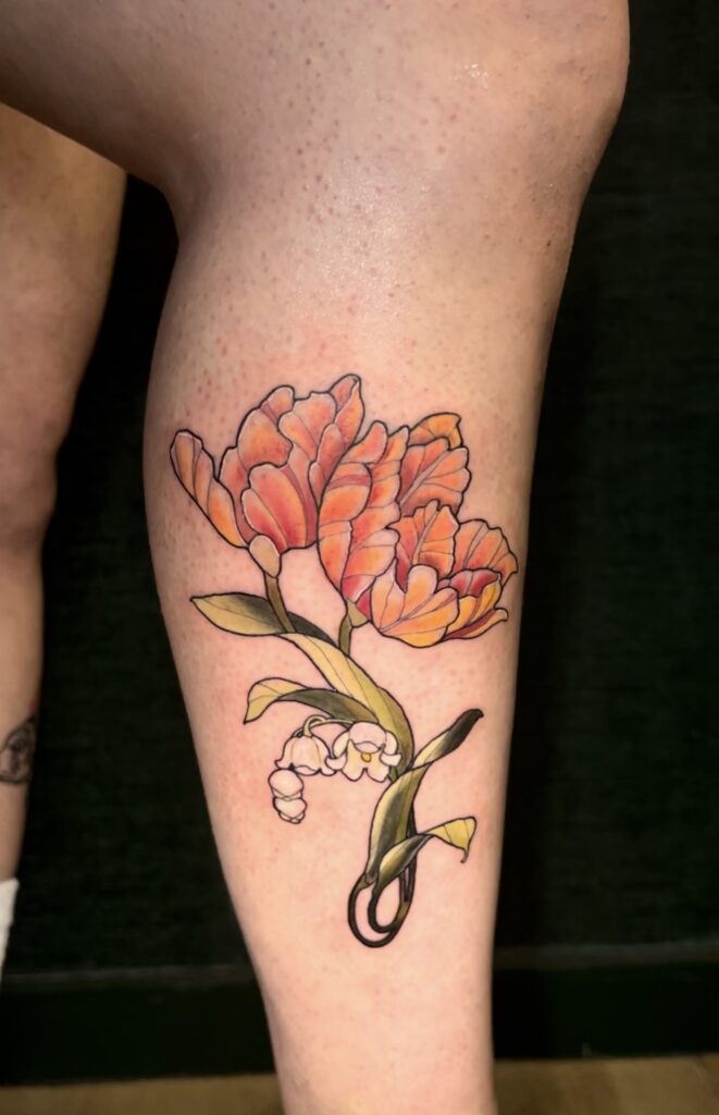 Een levendige tattoo op een onderbeen van oranje-rode tulpen gecombineerd met een takje witte Lelietjes-van-dalen. Het ontwerp heeft een zachte kleurovergang en strakke zwarte outlines. Gezet door Maeve van Maeve Sanaa op de Sint-Niklaas Tattoo Expo.