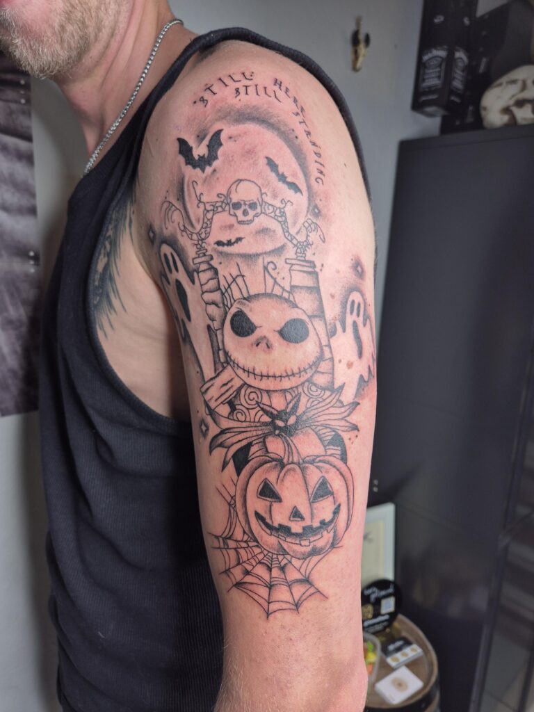 Een gedetailleerde zwart-inkt tattoo op een bovenarm geïnspireerd op 'The Nightmare Before Christmas'. Het ontwerp bevat Jack Skellington, een pompoen, spoken, vleermuizen en een volle maan, met de tekst 'Still here, still standing'. Gezet door Charis van Tattoo Charis op de Sint-Niklaas Tattoo Expo.