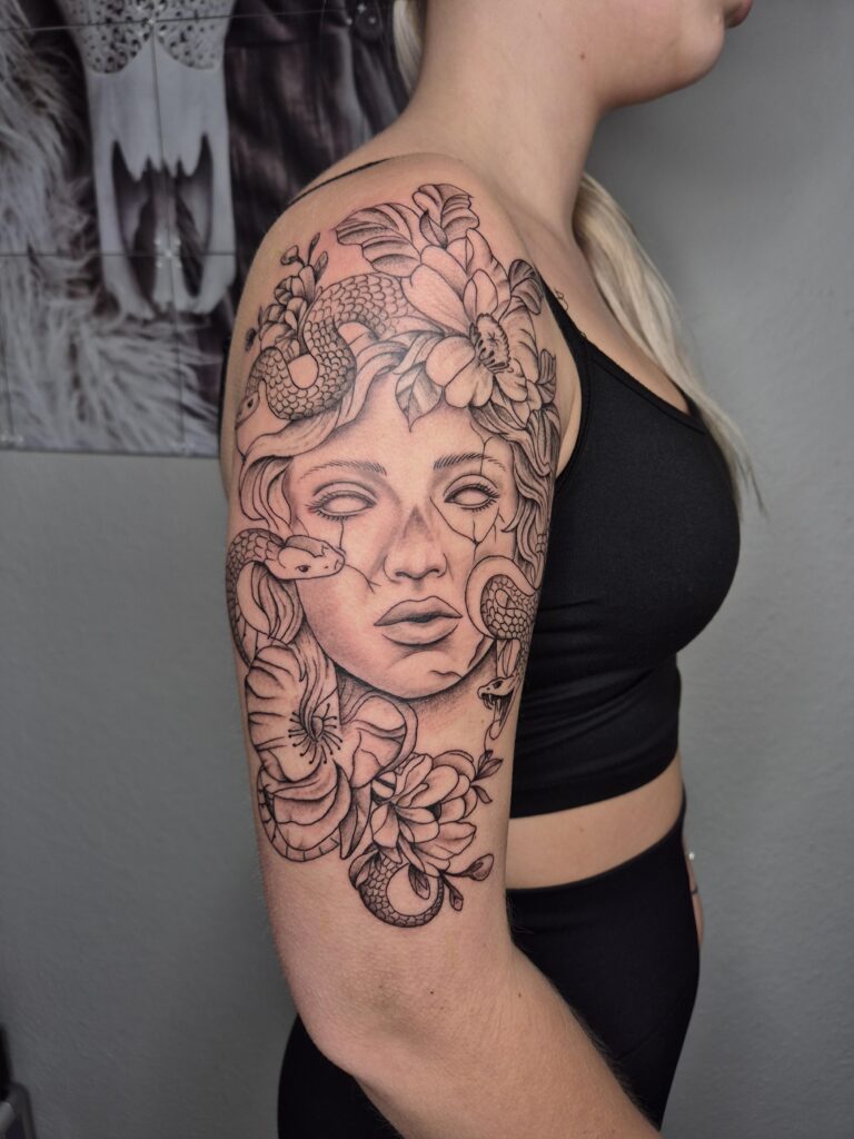 Een indrukwekkende black and grey tattoo op een bovenarm van een vrouwelijk gezicht (Medusa-stijl) omringd door slangen en bloemen. Het portret heeft een gebarsten stenen effect en witte ogen zonder pupillen. Gezet door Charis van Tattoo Charis op de Sint-Niklaas Tattoo Expo.