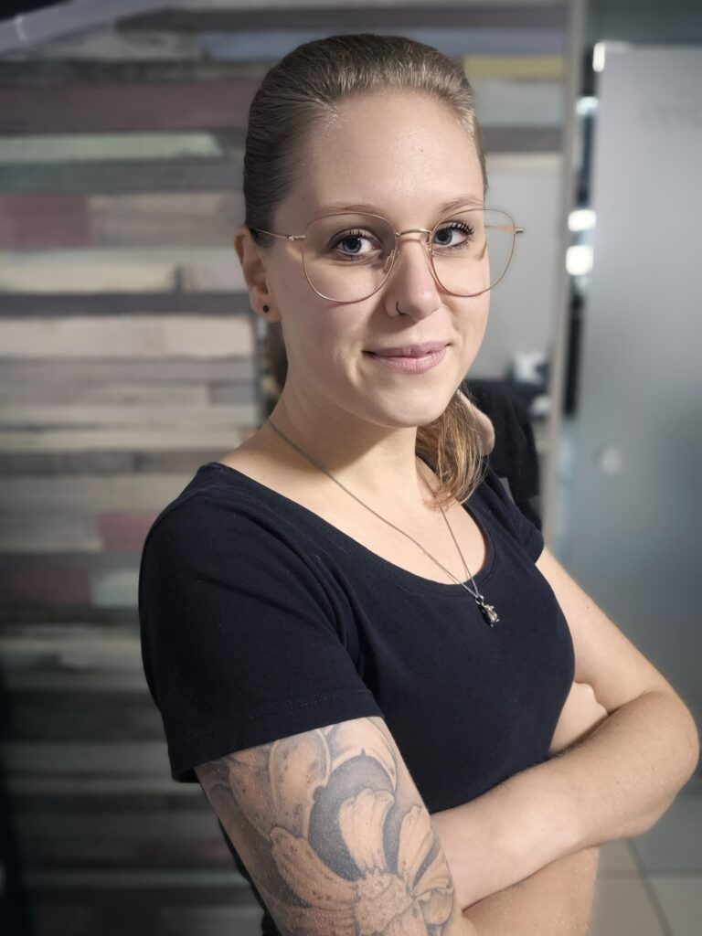 Portretfoto van tattoo-artiest Charis (Tattoo Charis) op de Sint-Niklaas Tattoo Expo. Ze draagt een bril met een goudkleurig montuur en een zwart T-shirt, met haar armen over elkaar geslagen waardoor haar eigen florale tattoos zichtbaar zijn.
