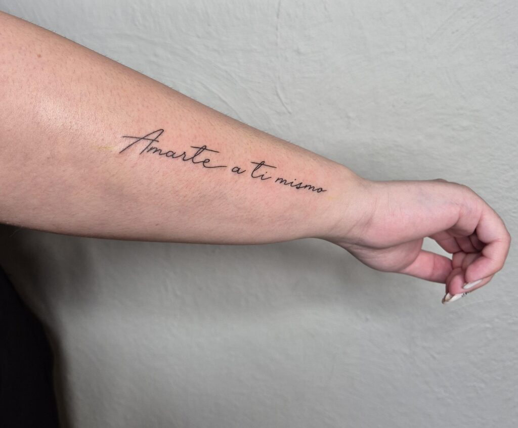 Een subtiele fineline tekst-tattoo op een onderarm met de Spaanse woorden 'Amarte a ti mismo' (houd van jezelf) in een elegant, handgeschreven lettertype. Werk van Charis (Tattoo Charis) op de Sint-Niklaas Tattoo Expo.
