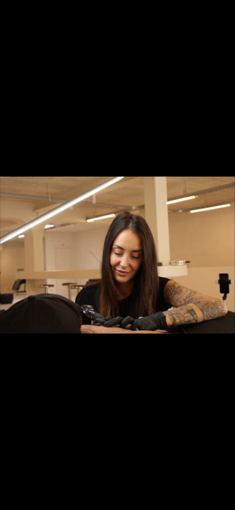 Een sfeervolle actiefoto van tattoo-artiest Marina (Serial Chillers) tijdens de Sint-Niklaas Tattoo Expo. Ze is geconcentreerd bezig met het zetten van een tatoeage. Marina heeft lang donker haar en haar eigen armen zijn voorzien van uitgebreide florale tattoos. Ze draagt zwarte handschoenen en werkt in een professioneel ingerichte studio-omgeving.