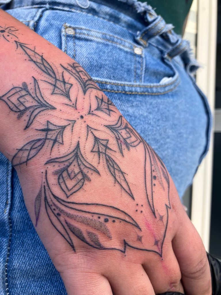 "Een verfijnde ornamentele handtattoo in handpoked stijl. Het ontwerp is een symmetrisch, bloemachtig patroon dat vanuit het midden over de handrug loopt, uitgevoerd met delicate lijnen en dotwork-accenten. Werk van Monica (Poked by Monique) op de Sint-Niklaas Tattoo Expo.