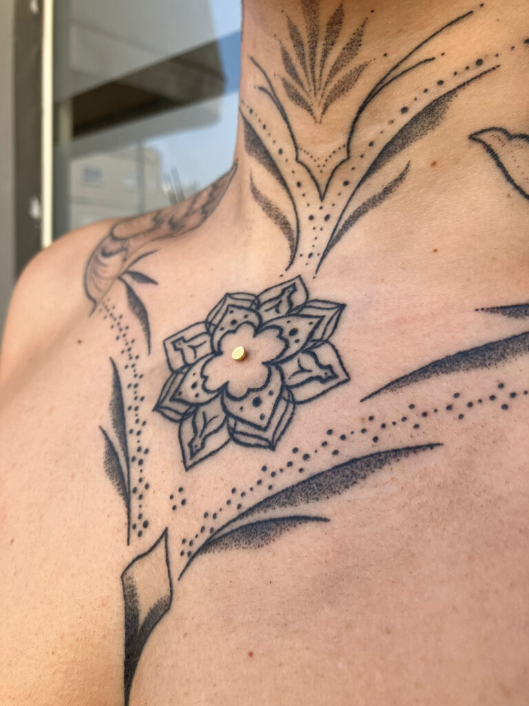 Een symmetrische ornamentele tattoo op de borst en hals, uitgevoerd in handpoked dotwork. In het midden van een klein mandala-bloemetje op het borstbeen is een goudkleurige dermal piercing geplaatst. Gezet door Monica (Poked by Monique) op de Sint-Niklaas Tattoo Expo.
